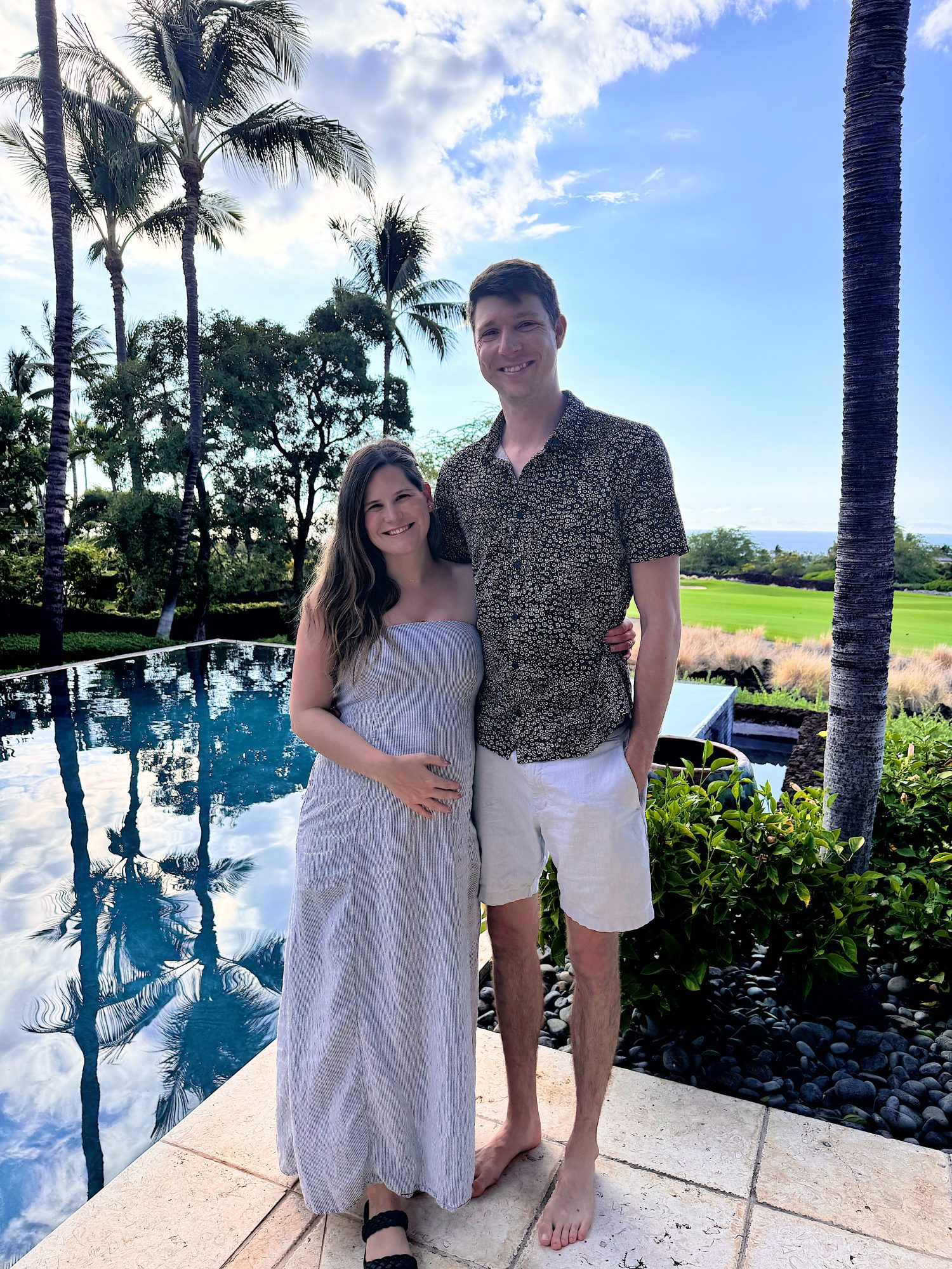 A couple stands side by side by a pool, surrounded by palm trees and greenery, with a blue sky and open landscape in the background.