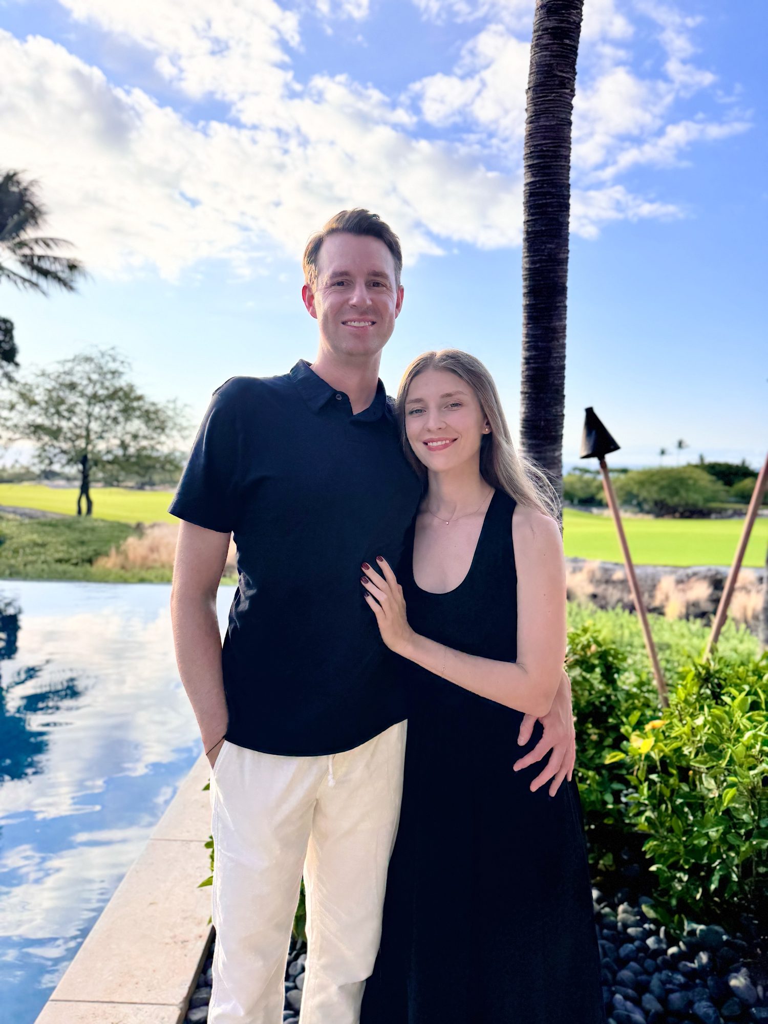 A man and woman stand together by a pool and lush greenery, with a palm tree and open sky in the background.