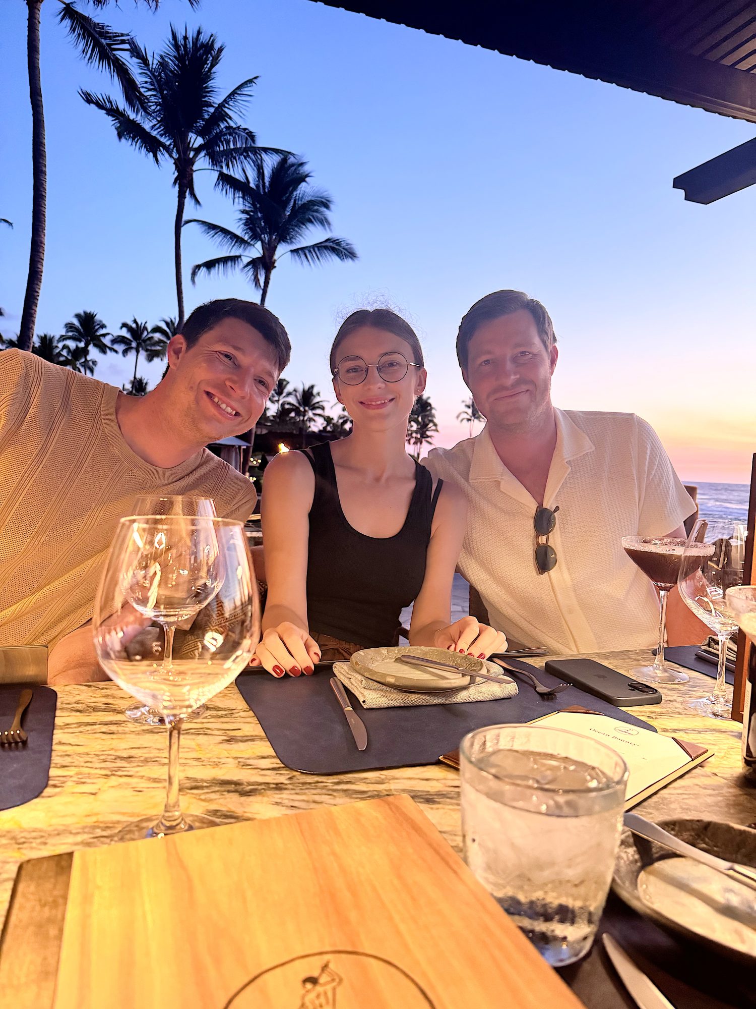 Three people sit at an outdoor restaurant table at sunset, with drinks, menus, and palm trees visible in the background.