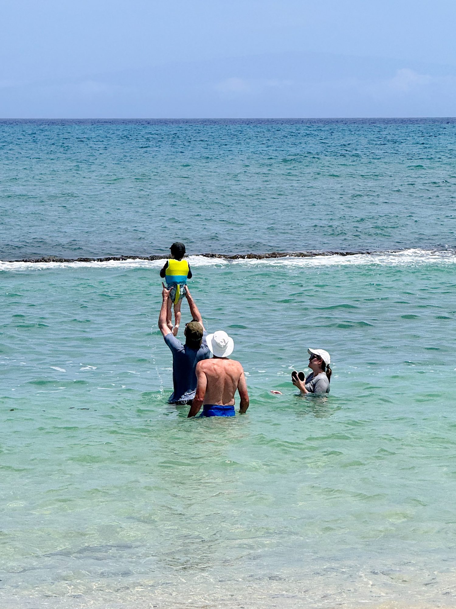 Three adults and a child in swimwear stand in shallow ocean water; one adult lifts the child in a life vest while another takes a photo.
