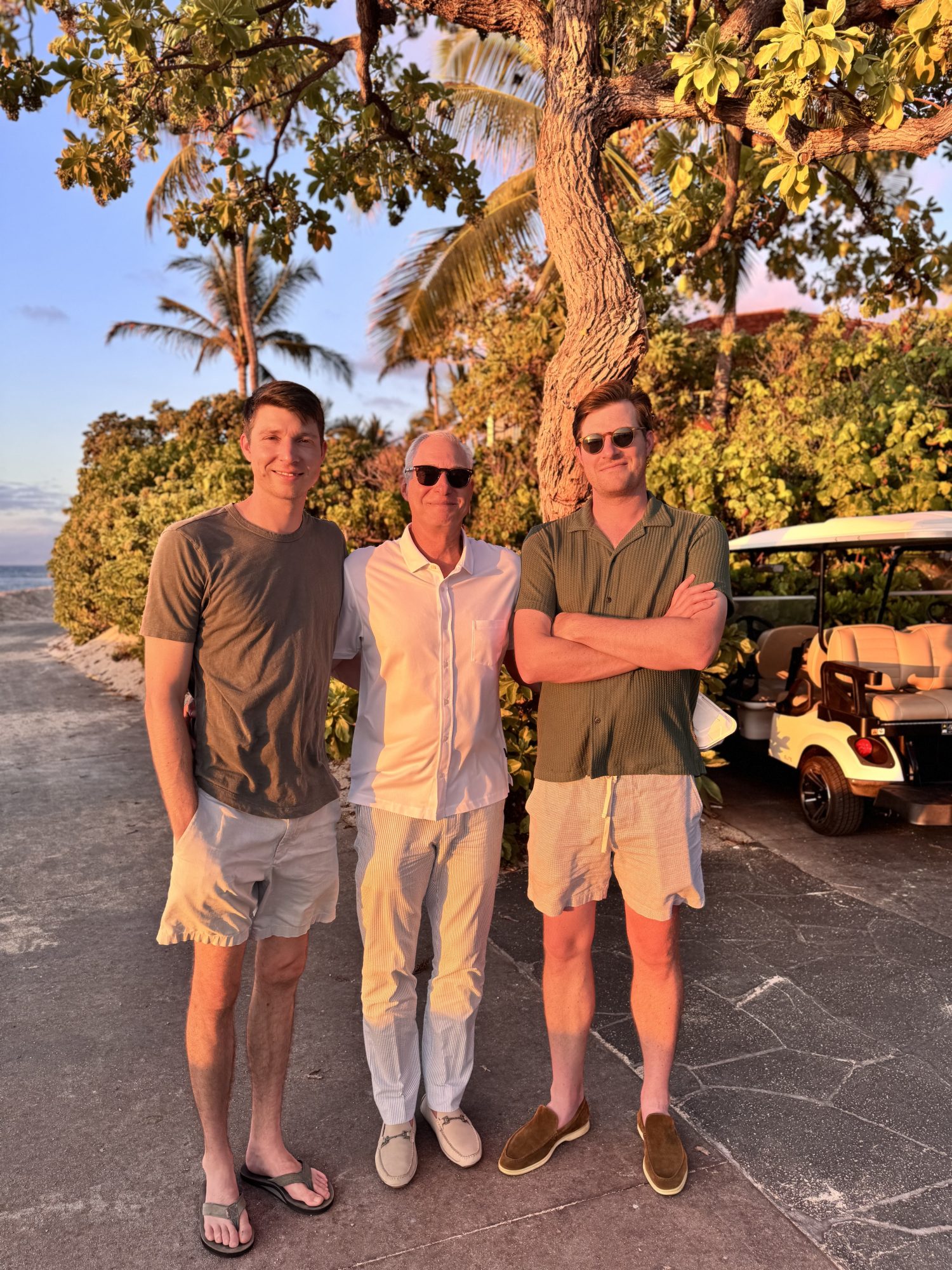 Three men stand outdoors on a sunny path near a golf cart and tropical trees, wearing summer clothes and sunglasses, with the beach visible in the background.
