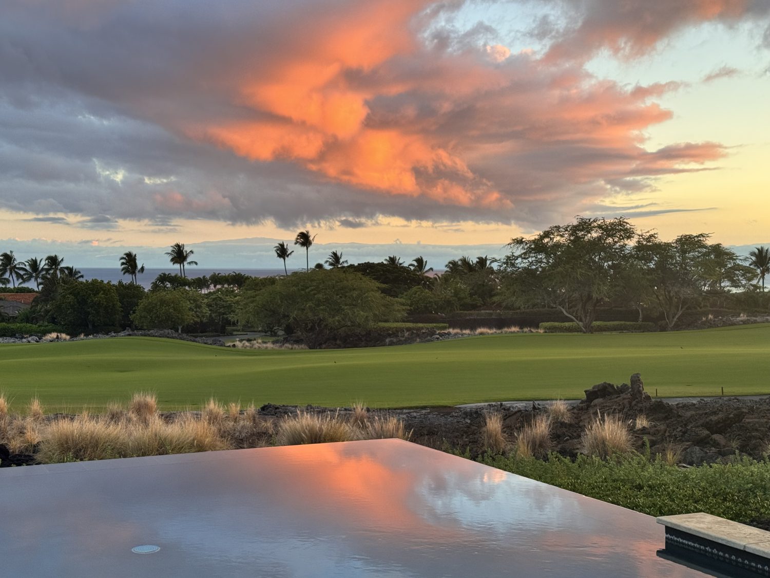 A vibrant sunset sky with orange clouds is reflected in a pool overlooking a golf course, with palm trees and greenery in the background.