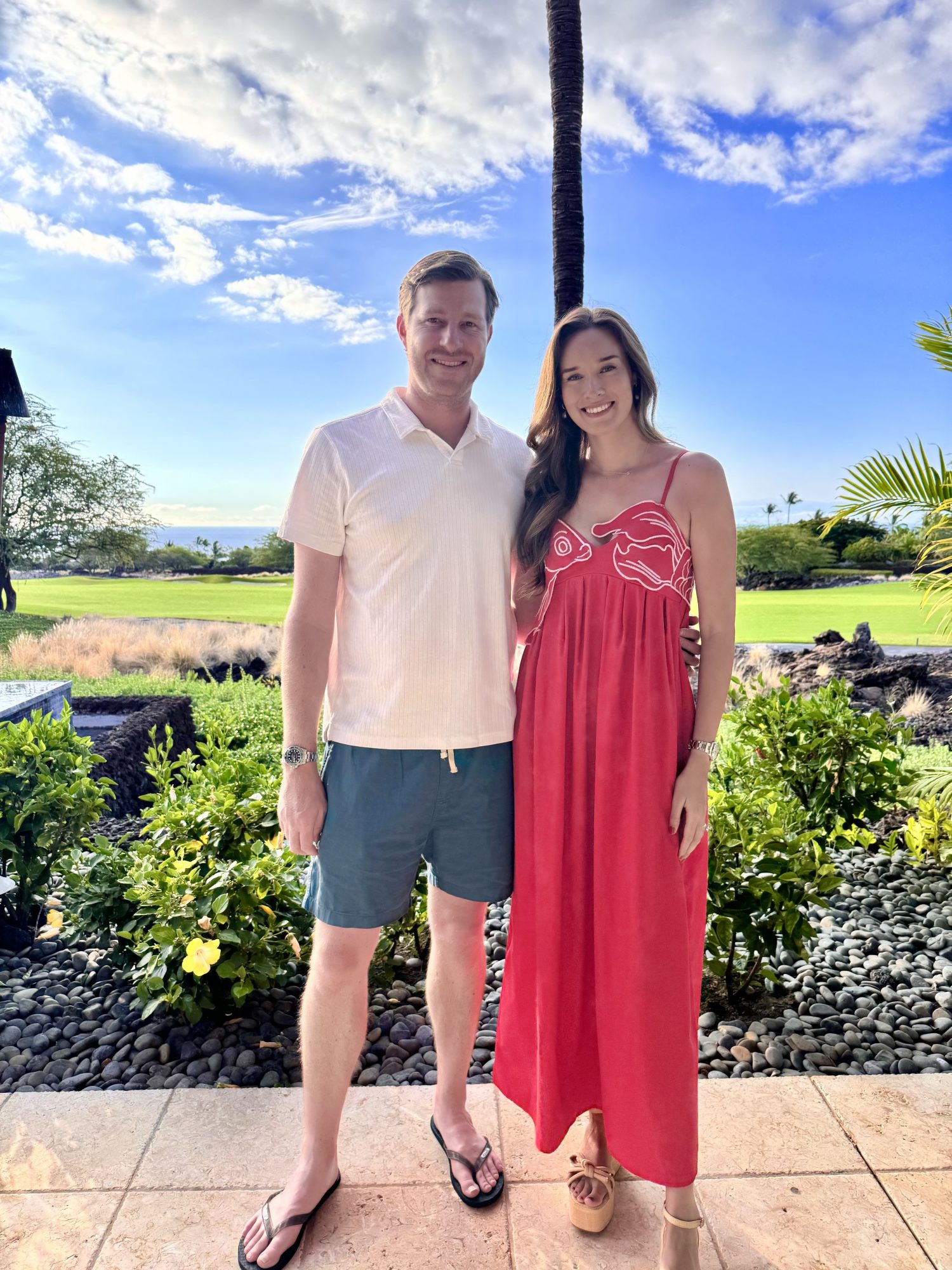 A man in a white shirt and shorts stands next to a woman in a red dress outside on a patio, with greenery and blue sky in the background.