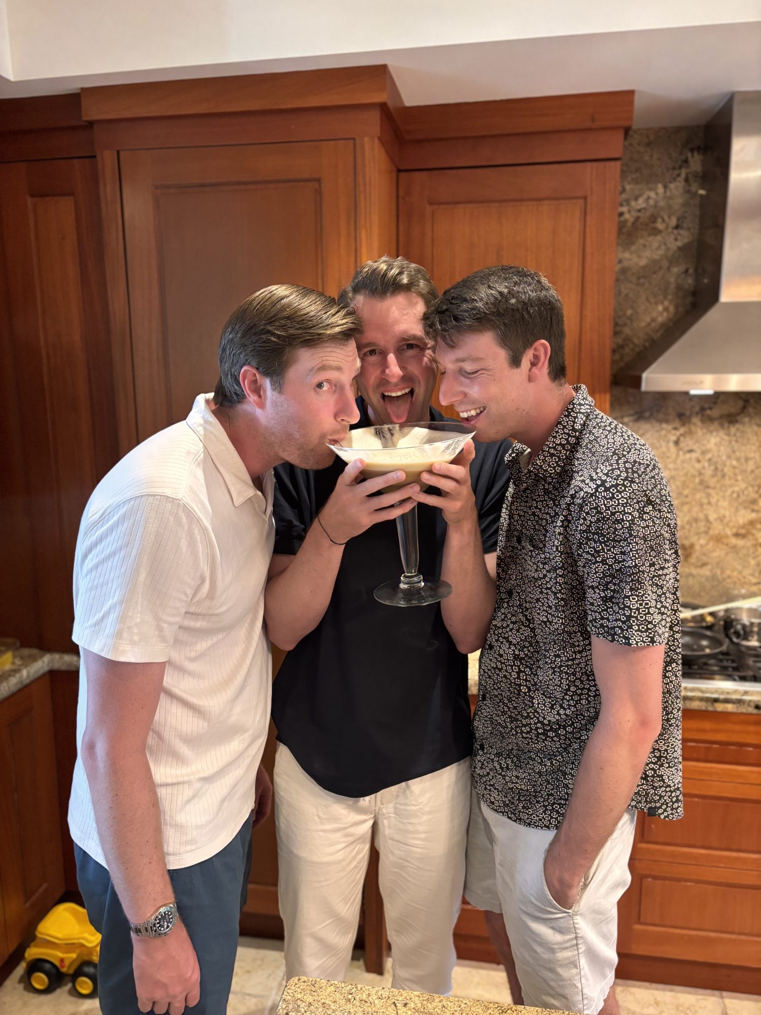 Three men are sharing a large margarita glass with straws in a kitchen, standing close together and smiling.