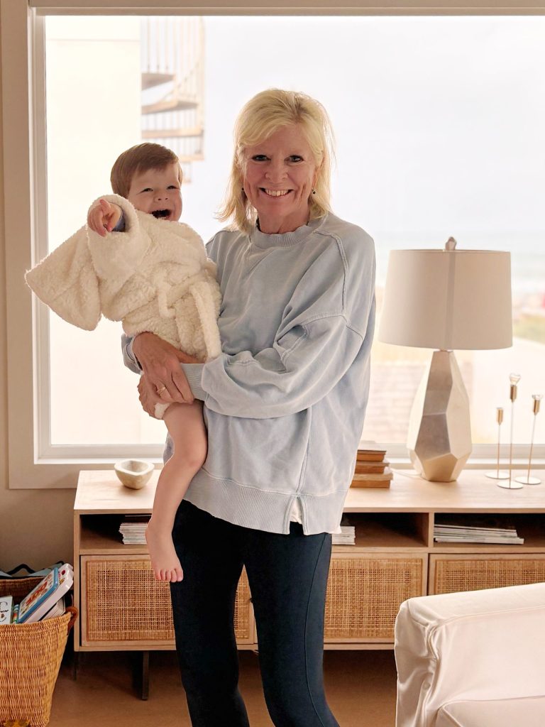 A woman in a light sweatshirt holds a small child wearing a fluffy robe, standing in a bright living room with a window and a lamp in the background.