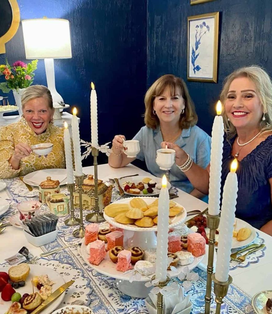 Three women sit at a decorated table with candles, pastries, and tea, smiling and holding teacups in a blue-walled room with framed art.