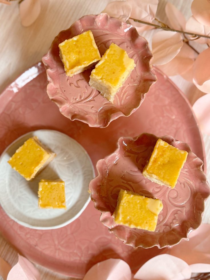 Four square pieces of yellow cake are displayed on three decorative plates, one white and two pink, with pale pink leaves in the background.