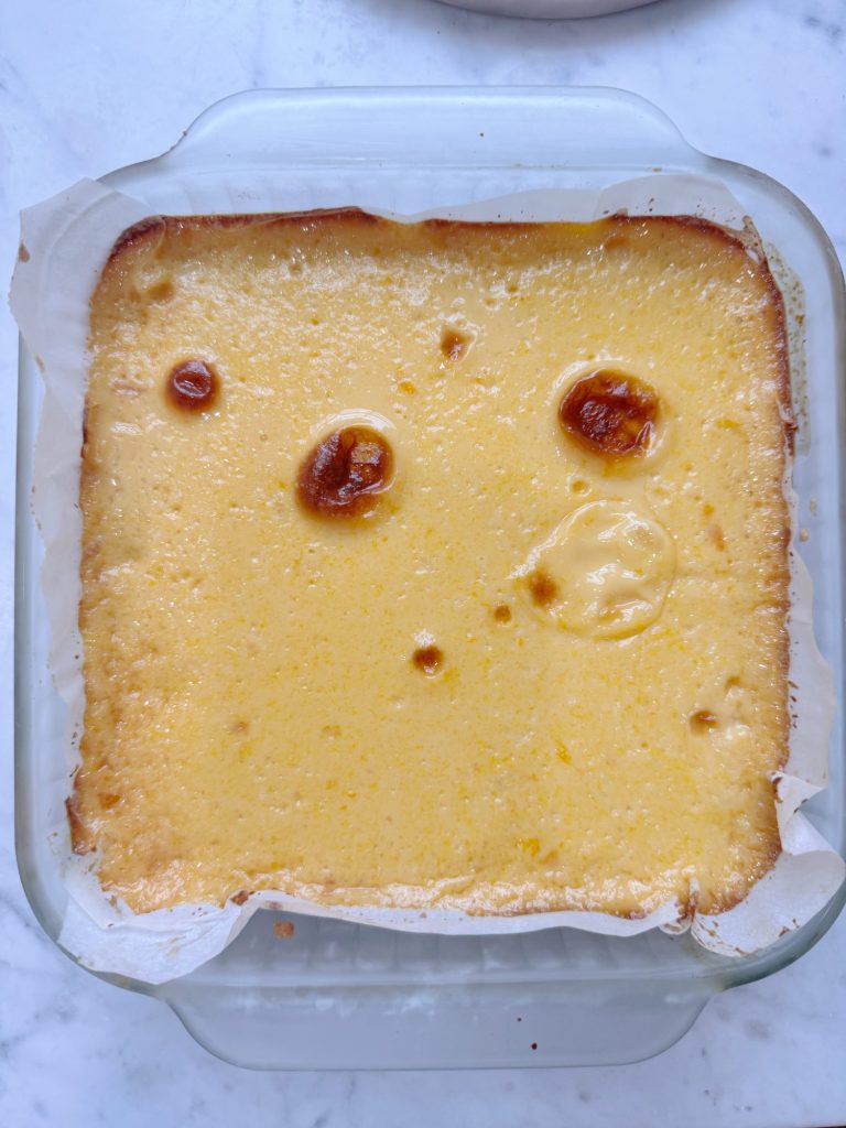 A square glass baking dish filled with a yellow baked custard or cheesecake, with browned spots and lined with parchment paper.