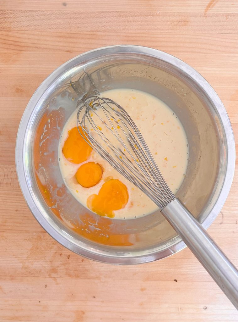 A metal mixing bowl with three cracked eggs and batter, with a whisk resting inside, sits on a wooden surface.