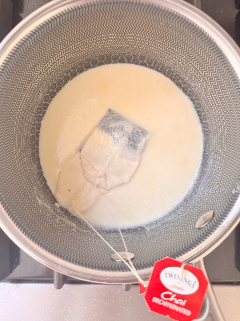 A chai tea bag steeps in a saucepan of milk on a stovetop. The tea tag labeled "Twinings Chai Decaffeinated" hangs outside the pot.