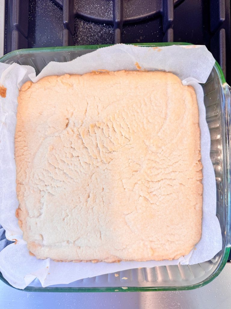 A square glass baking dish lined with parchment paper holds a single layer of baked shortbread with a lightly golden surface and visible fork marks.