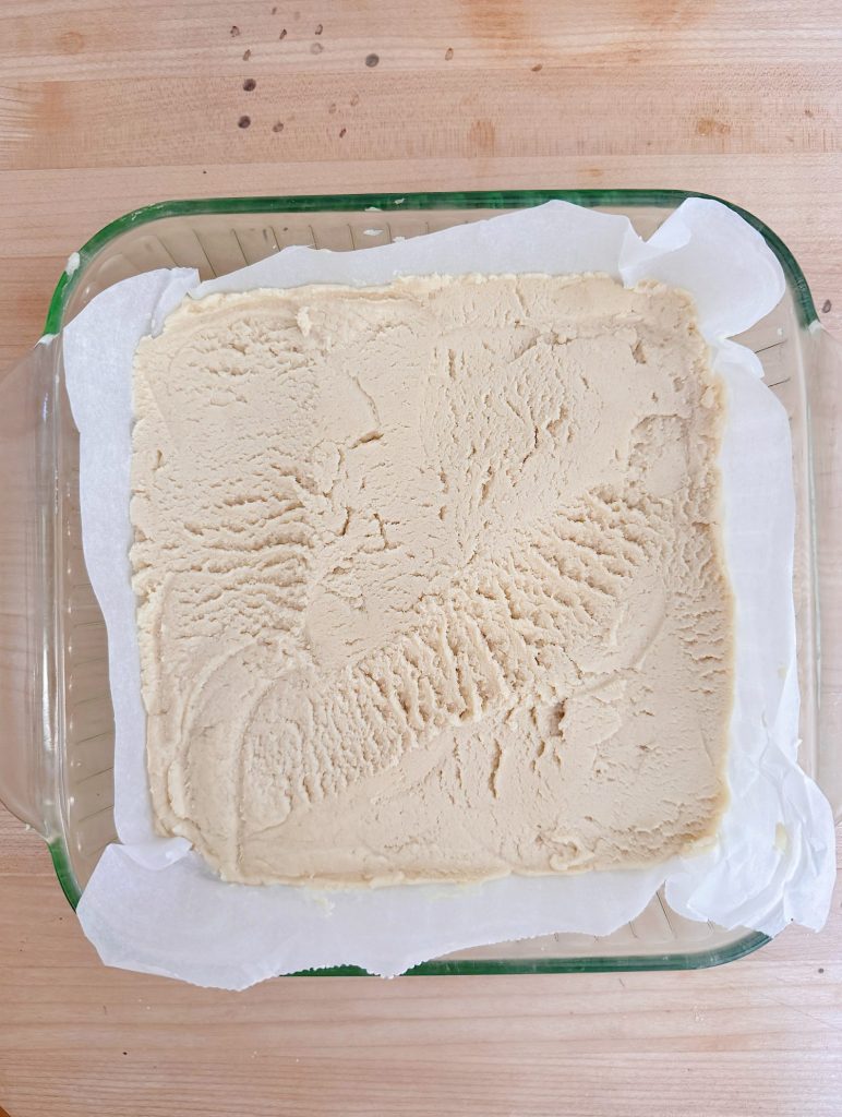 A glass baking dish lined with parchment paper contains an even layer of unbaked dough, spread flat with visible utensil marks on the surface.
