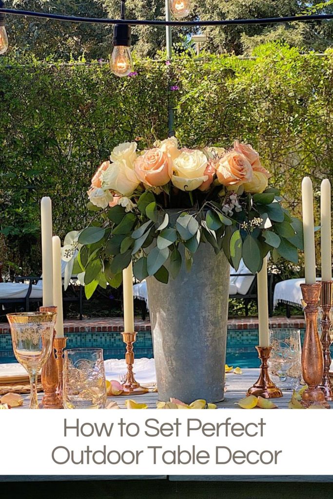 A metal vase with pink and white roses sits on an outdoor table set with candles, glasses, and copper accents in front of greenery and string lights.