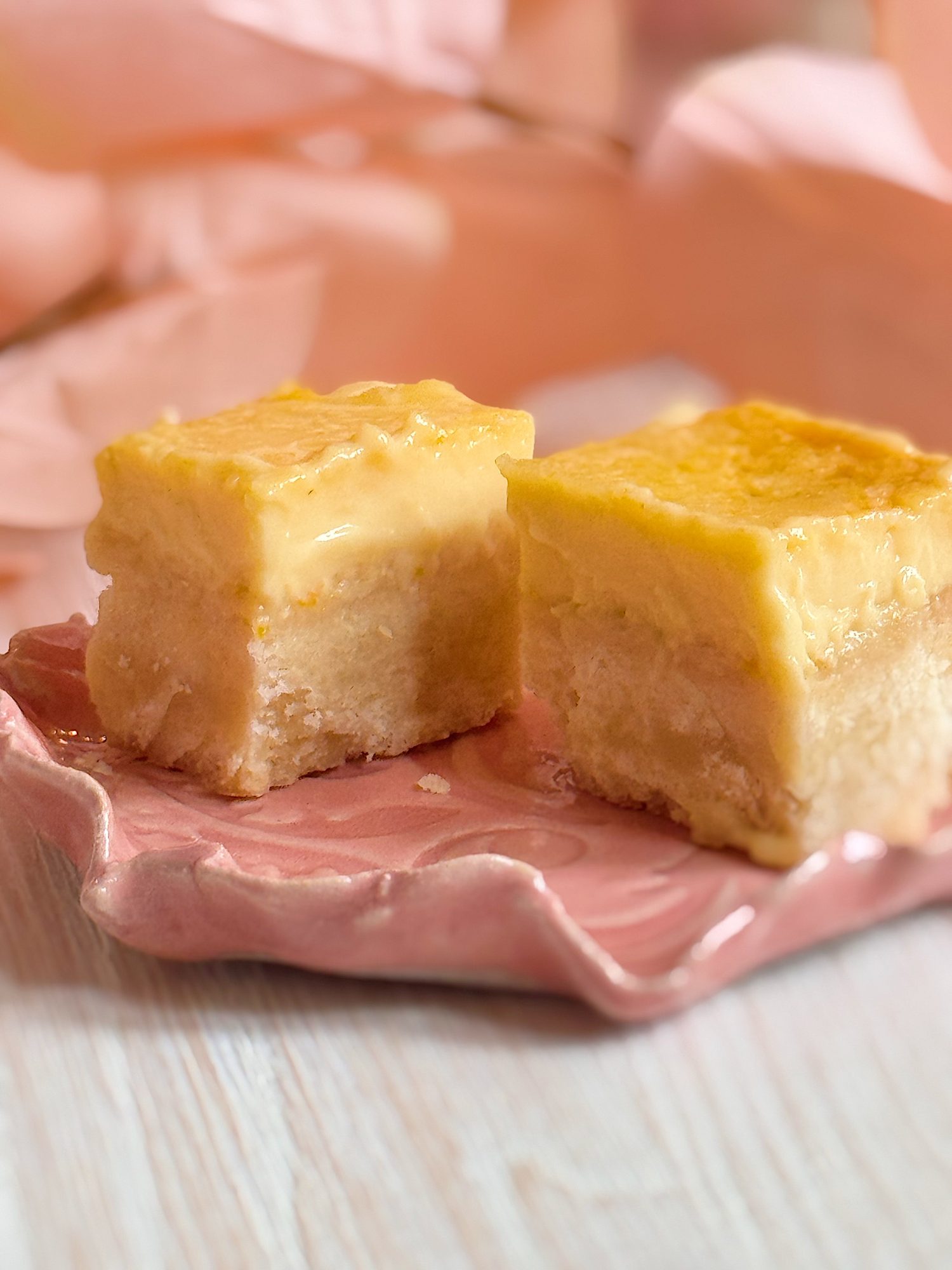 Two square slices of layered custard cake are placed on a pink ceramic plate, with a light wooden surface and blurred background.