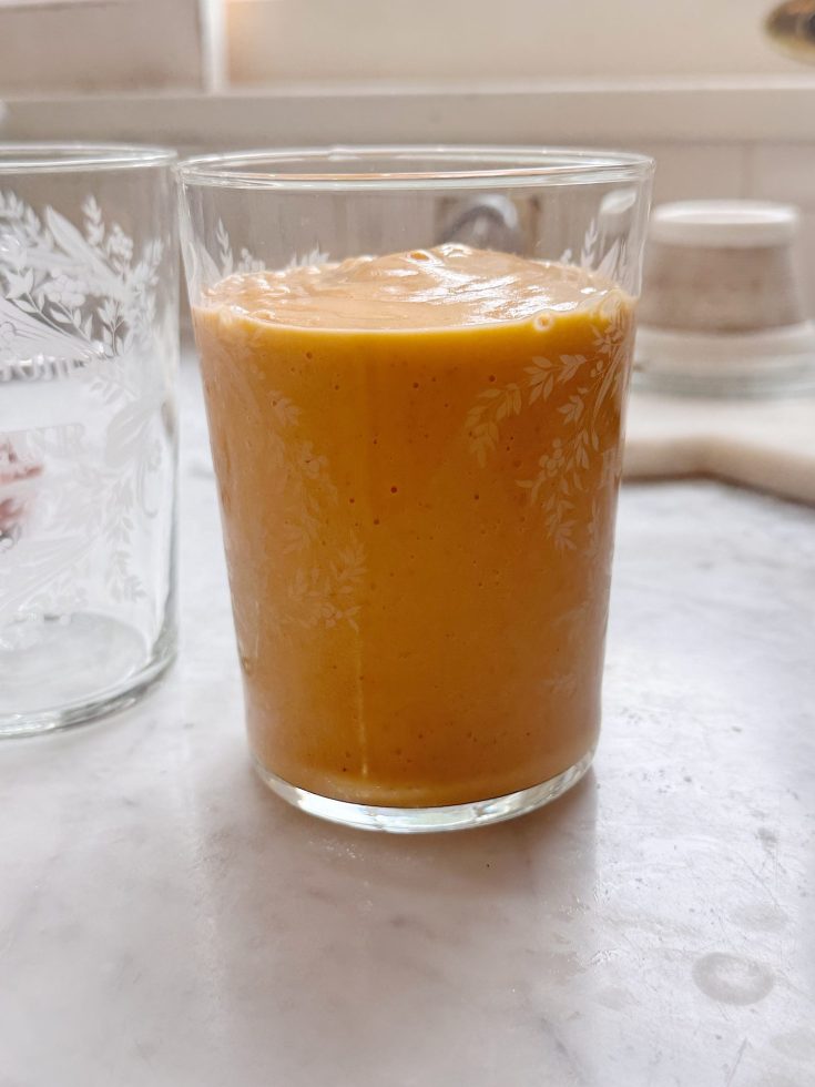 A clear glass filled with a thick, orange smoothie sits on a marble countertop next to an empty glass.