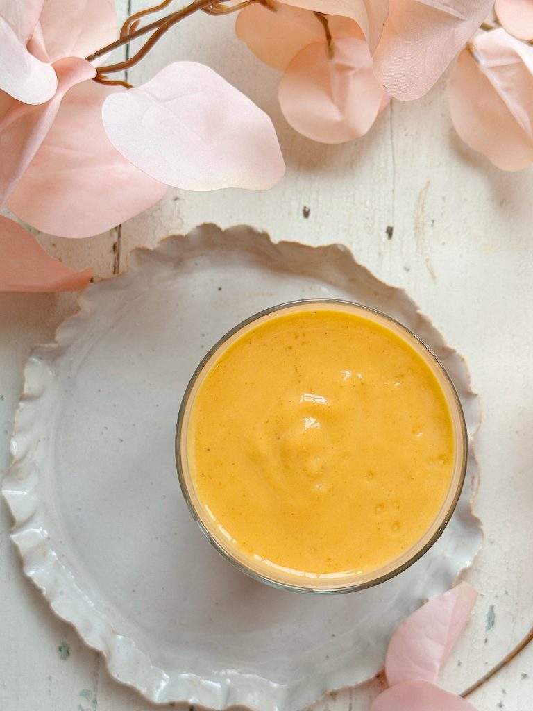 A glass of thick orange smoothie sits on a white ceramic plate, surrounded by pale pink artificial leaves on a white wooden surface.