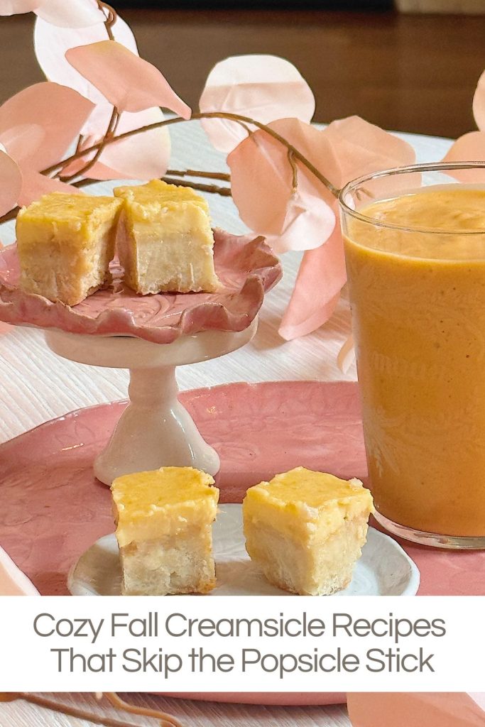 A glass of orange creamsicle smoothie sits next to small square dessert bars on a pink and white plate with pink foliage in the background.