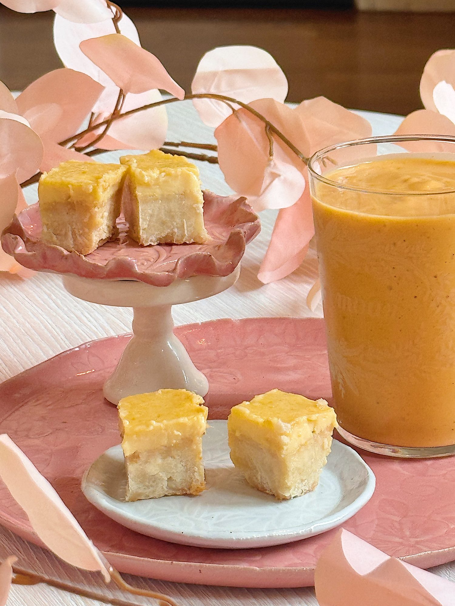 Small yellow-topped dessert squares are displayed on pink and white plates next to a glass of orange smoothie, with pink decorative leaves in the background.