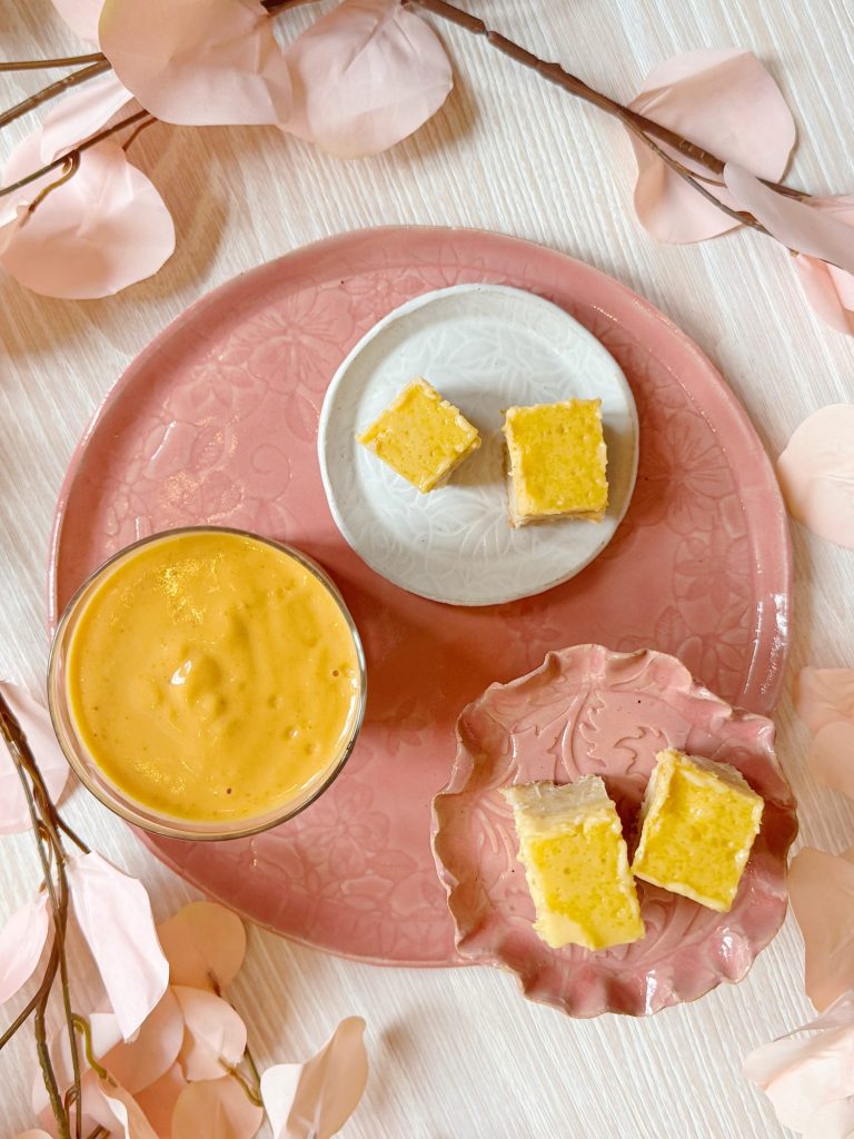A pink plate with a glass of orange smoothie and three small yellow square desserts on separate dishes, surrounded by pink artificial flowers.