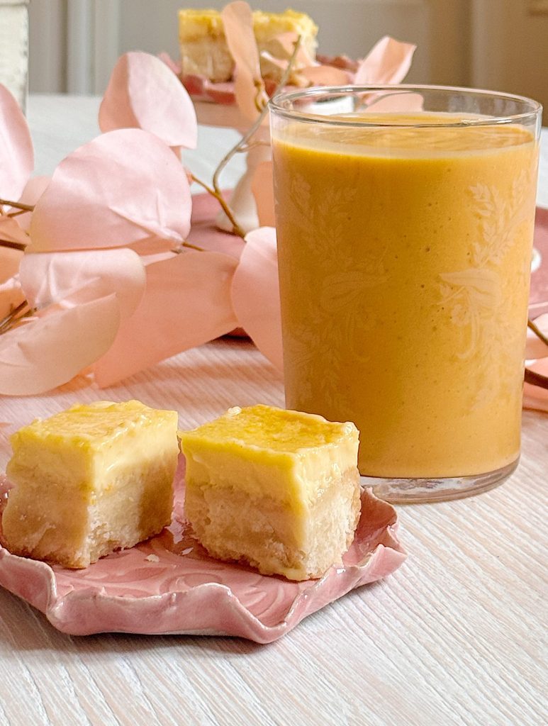 A glass of orange smoothie next to two small lemon bars on a pink ceramic plate, with pink decorative leaves in the background.