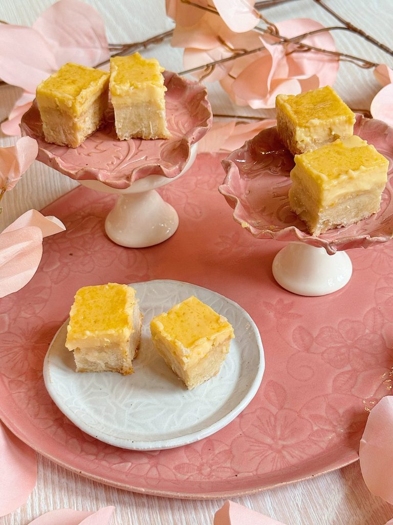 Six small square pieces of cake are displayed on two pink pedestal plates and one white plate, all set on a pink tray with floral designs and pink decorative leaves.