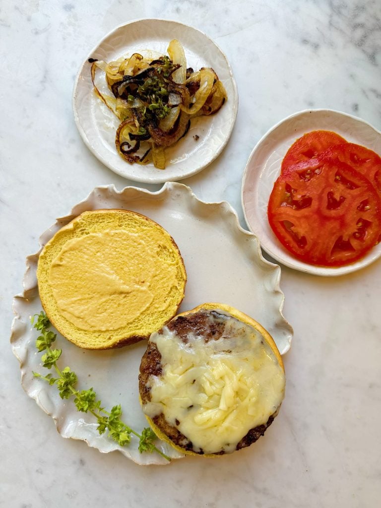An open cheeseburger with melted cheese on a bun, next to leafy herbs, sliced tomatoes, and a plate of grilled onions and peppers on a marble surface.