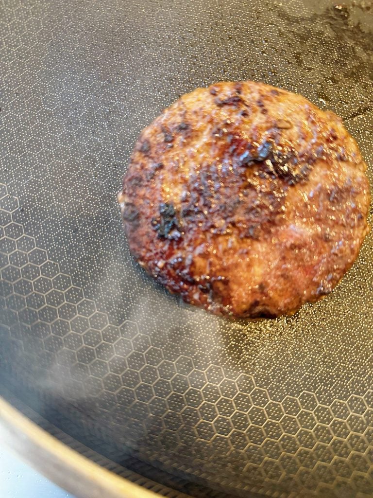 A cooked hamburger patty sits in a frying pan with visible grill marks and some grease.