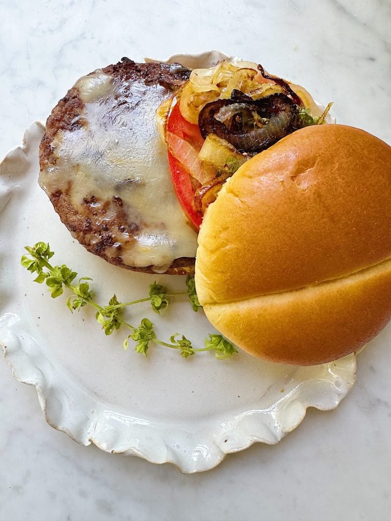 Open cheeseburger with grilled onions, tomato, and melted cheese on a white plate, accompanied by a sprig of herbs on a marble surface.