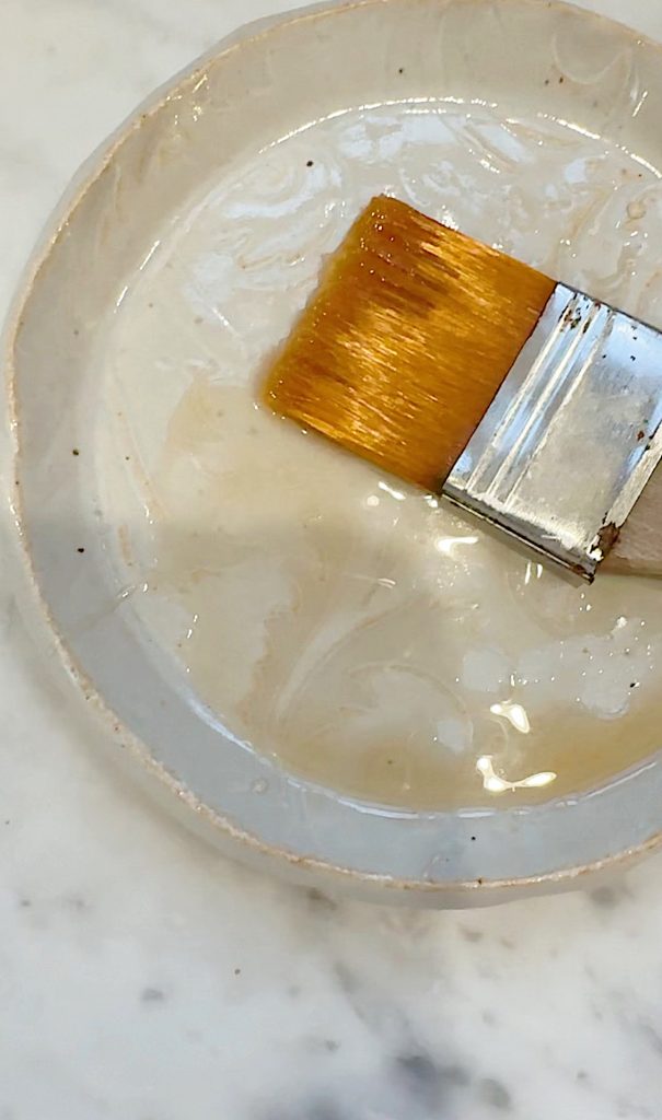 A paintbrush with light brown bristles rests in a shallow white dish filled with clear liquid, placed on a light-colored marble surface.