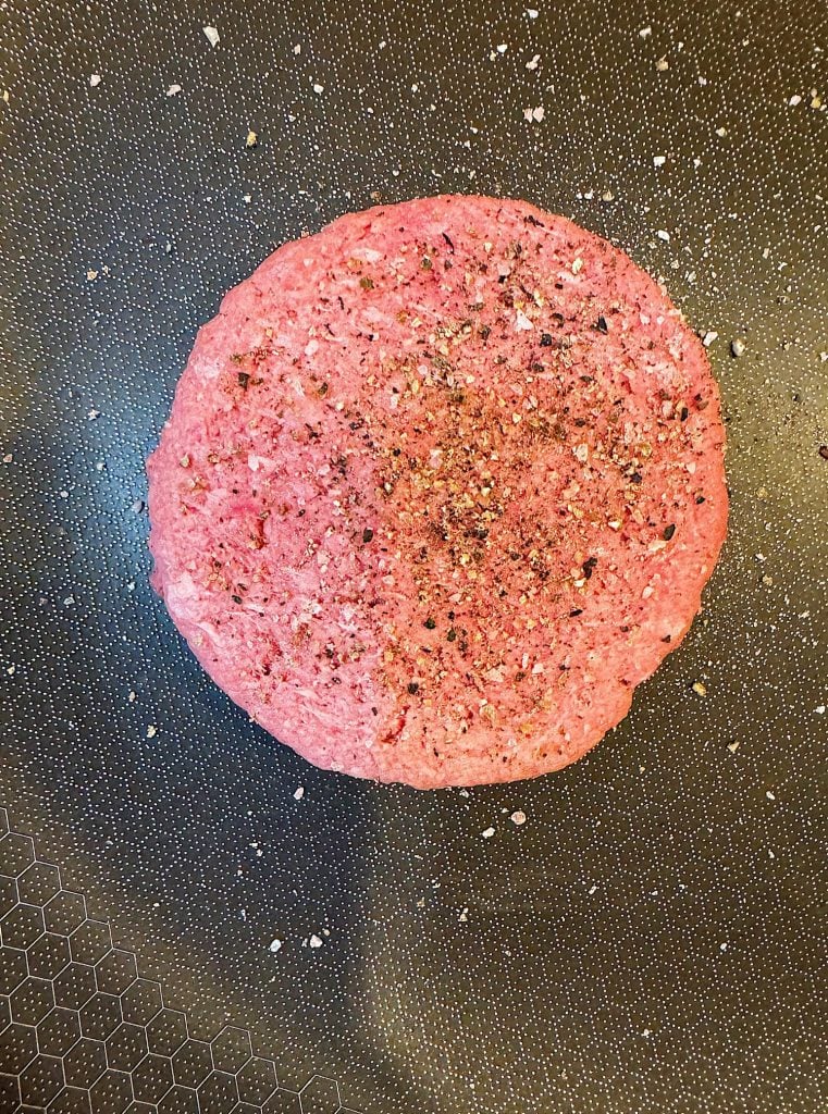 A raw seasoned burger patty sits in a nonstick frying pan, ready to be cooked.