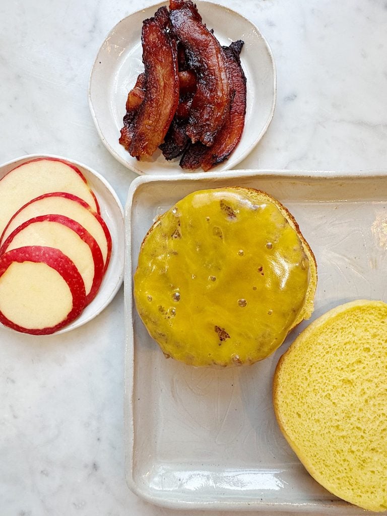 A cheeseburger bun with melted cheese, a small plate of crispy bacon strips, and another plate with sliced red apples on a marble surface.