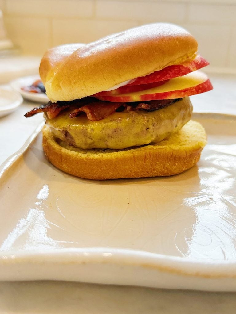 A cheeseburger on a square ceramic plate, topped with bacon, tomato slices, and apple slices, on a lightly toasted bun.
