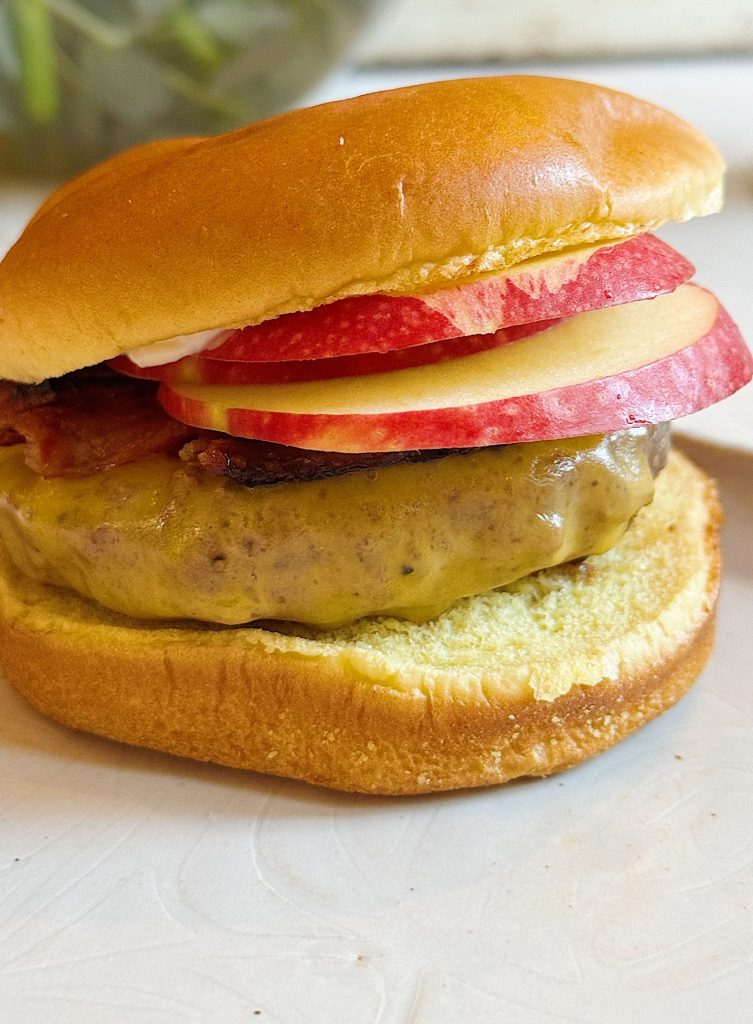 Cheeseburger on a bun topped with bacon, apple slices, and mayonnaise, served on a white plate.
