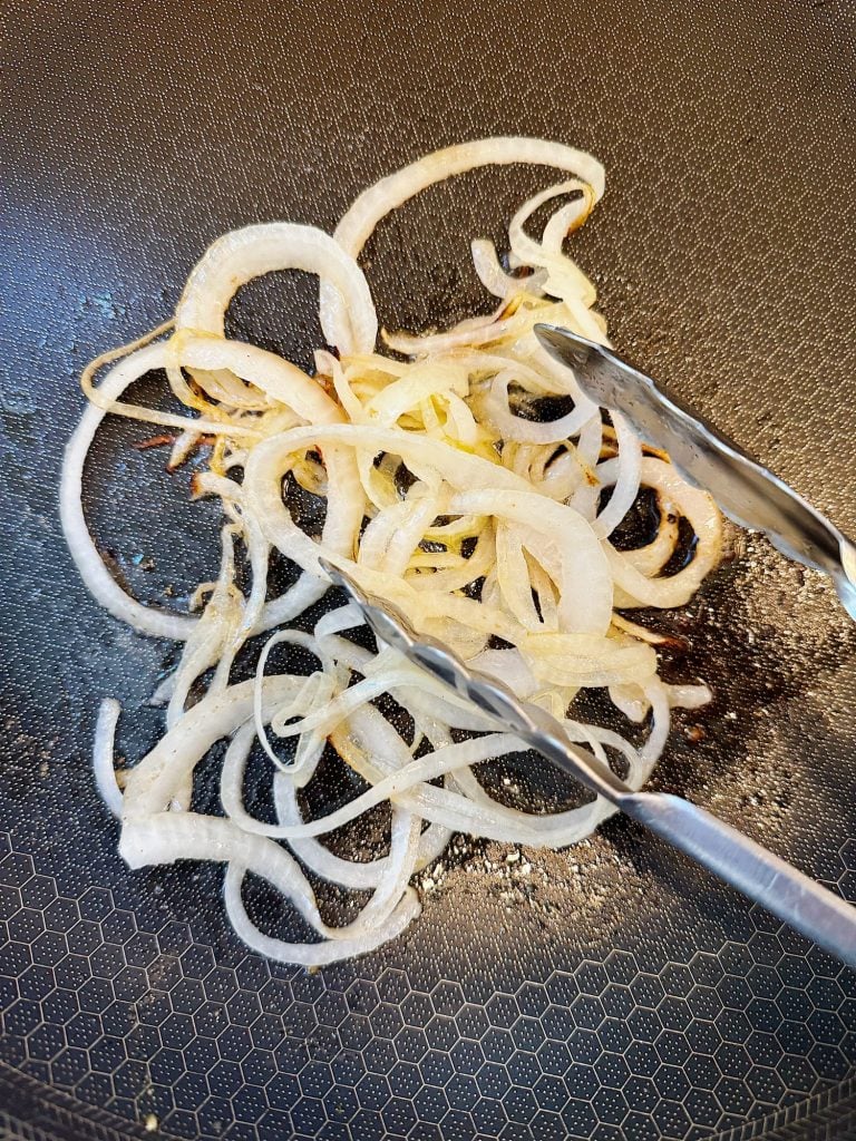 Sliced onions being sautéed in a pan with metal tongs visible.