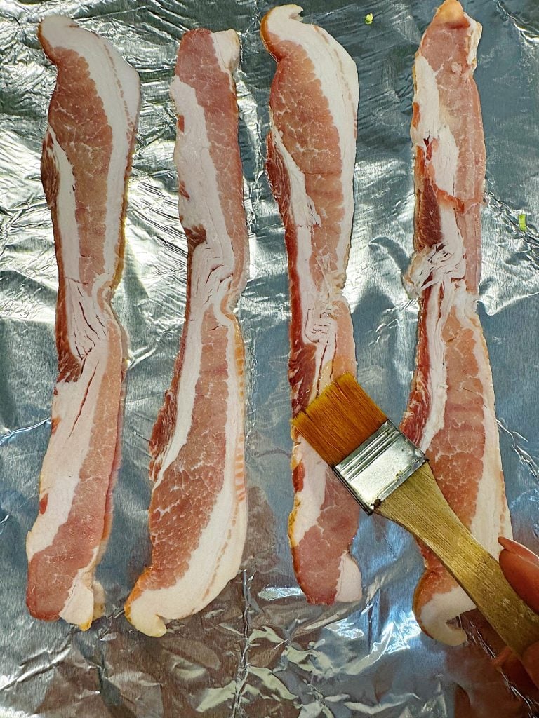 Four raw bacon strips on foil with a hand brushing one strip using a wooden pastry brush.