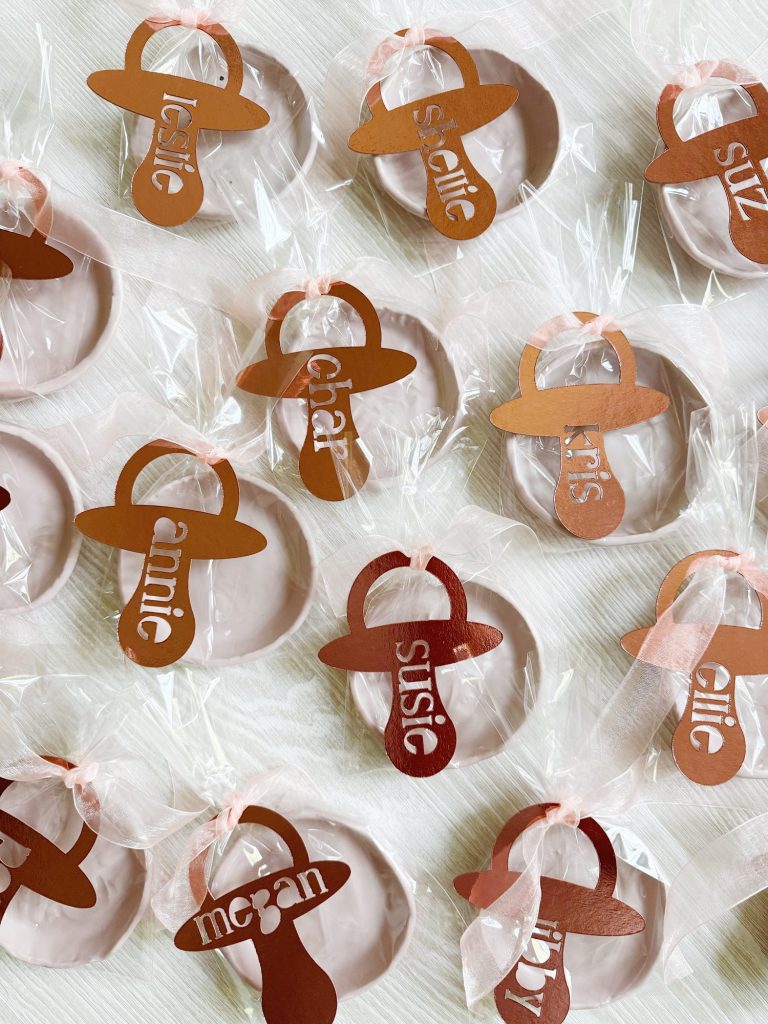Multiple round cookies wrapped in clear plastic, each topped with a metallic pacifier-shaped tag labeled with a different name in white lowercase letters.