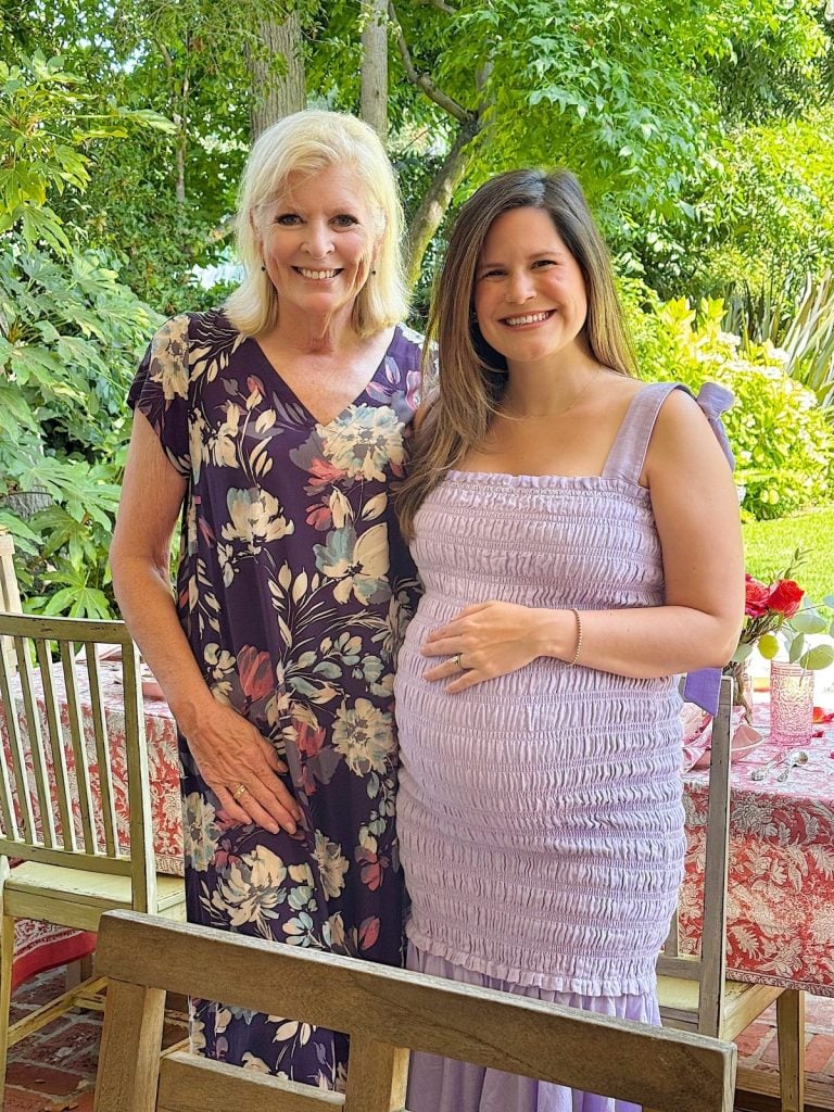 Two women stand side by side outdoors, smiling at the camera. One woman wears a floral dress and the other, who appears pregnant, wears a light purple dress. Greenery is visible in the background.