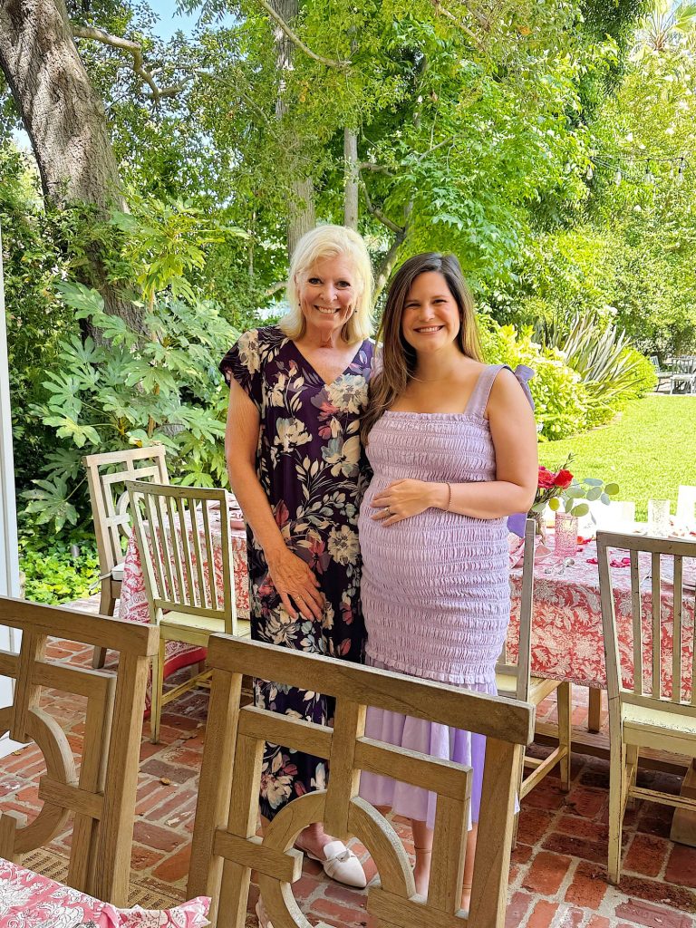 Two women stand and smile in an outdoor garden setting with a brick patio and a table set for a meal. One woman is touching her pregnant belly.