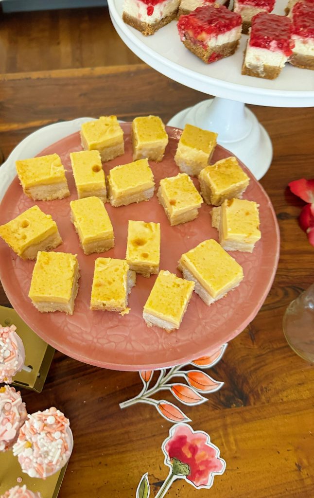 A pink plate holds several square pieces of yellow dessert bars. Other assorted desserts and paper flower decorations are visible on the wooden table.