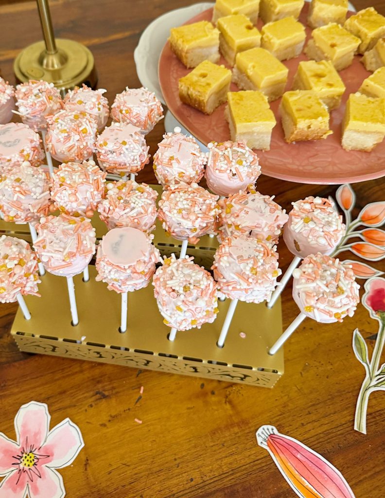 A display of pink and white sprinkle-covered cake pops on a stand, next to a plate of yellow cake squares on a wooden table with floral decorations.