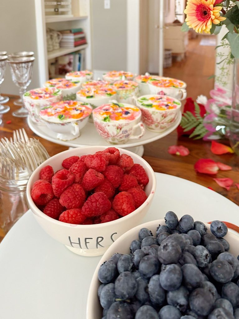 A bowl of raspberries, a bowl of blueberries, and a cake stand with decorated teacups are arranged on a table with flowers and glassware in the background.