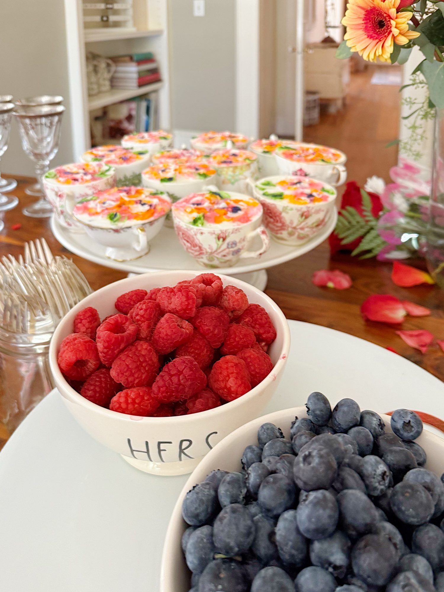 A bowl of raspberries, a bowl of blueberries, and a cake stand with decorated teacups are arranged on a table with flowers and glassware in the background.
