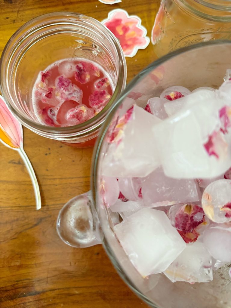 A glass with a pink drink and floating berries sits next to a bowl of ice cubes with flowers and berries inside, on a wooden surface with floral paper cutouts.