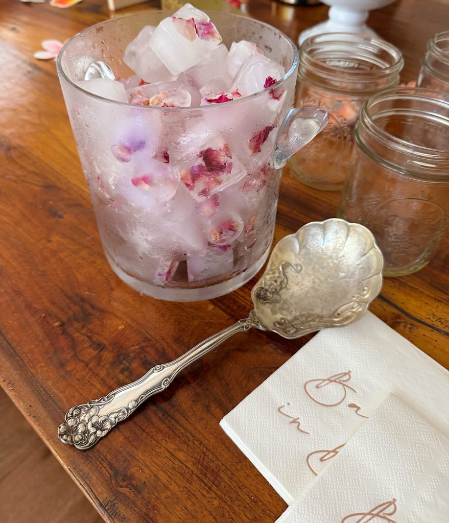 A glass mug filled with ice cubes containing rose petals sits on a wooden table beside an ornate silver spoon, napkins, and empty glass jars.
