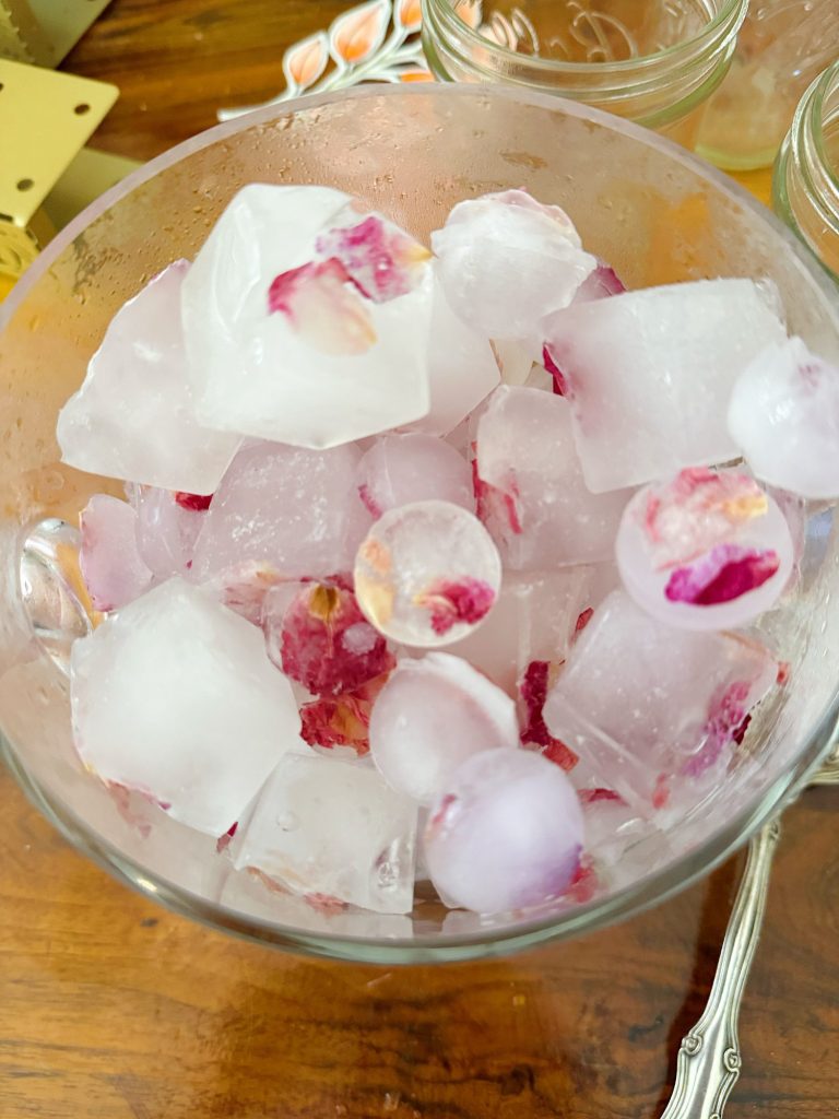 A glass bowl filled with ice cubes, some containing frozen flower petals, on a wooden surface.