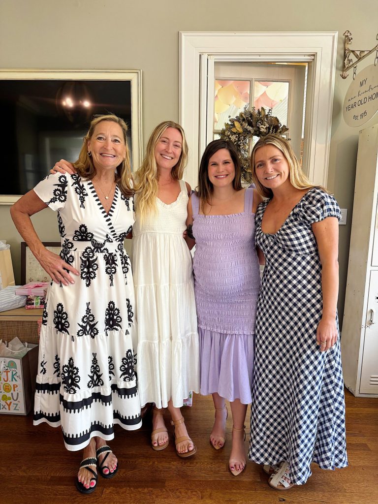 Four women stand side by side indoors, smiling at the camera, each wearing a different patterned dress.