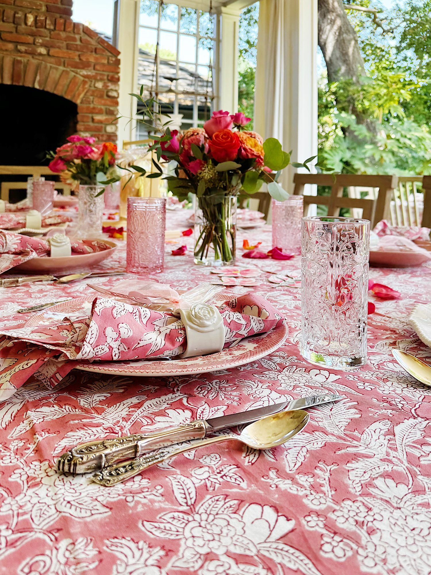 A table set for a meal with pink floral tablecloth, matching napkins, floral centerpieces, clear glasses, and silverware, in a bright room with a view of greenery outside.