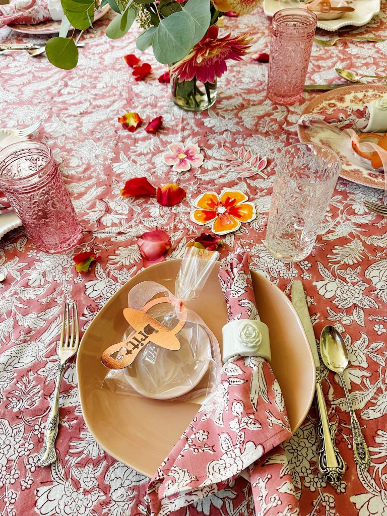 A table set with pink floral-patterned linens, peach plates, pink glassware, silver cutlery, floral decorations, and a place setting with a wrapped gift and name tag.