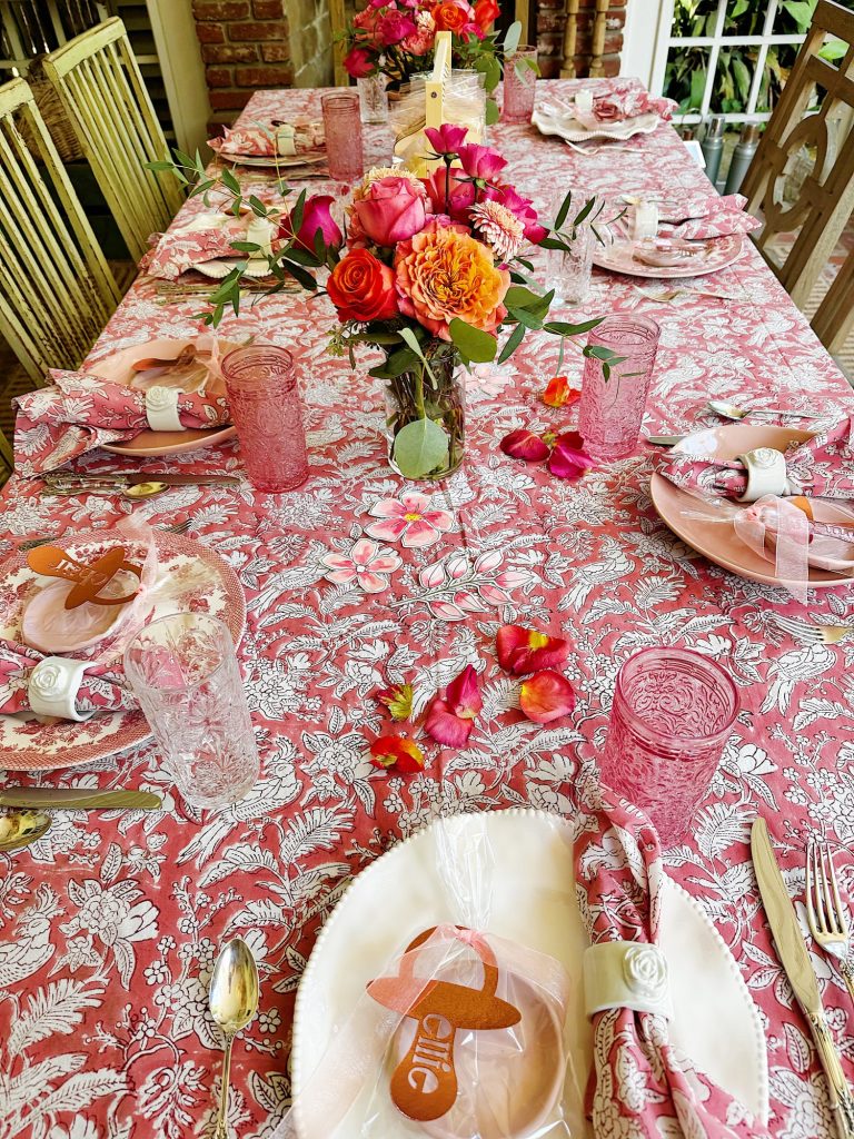 A table set for a meal with pink floral tablecloth, matching napkins, pink glasses, plates, floral centerpieces, and name-tagged cookies at each place setting.