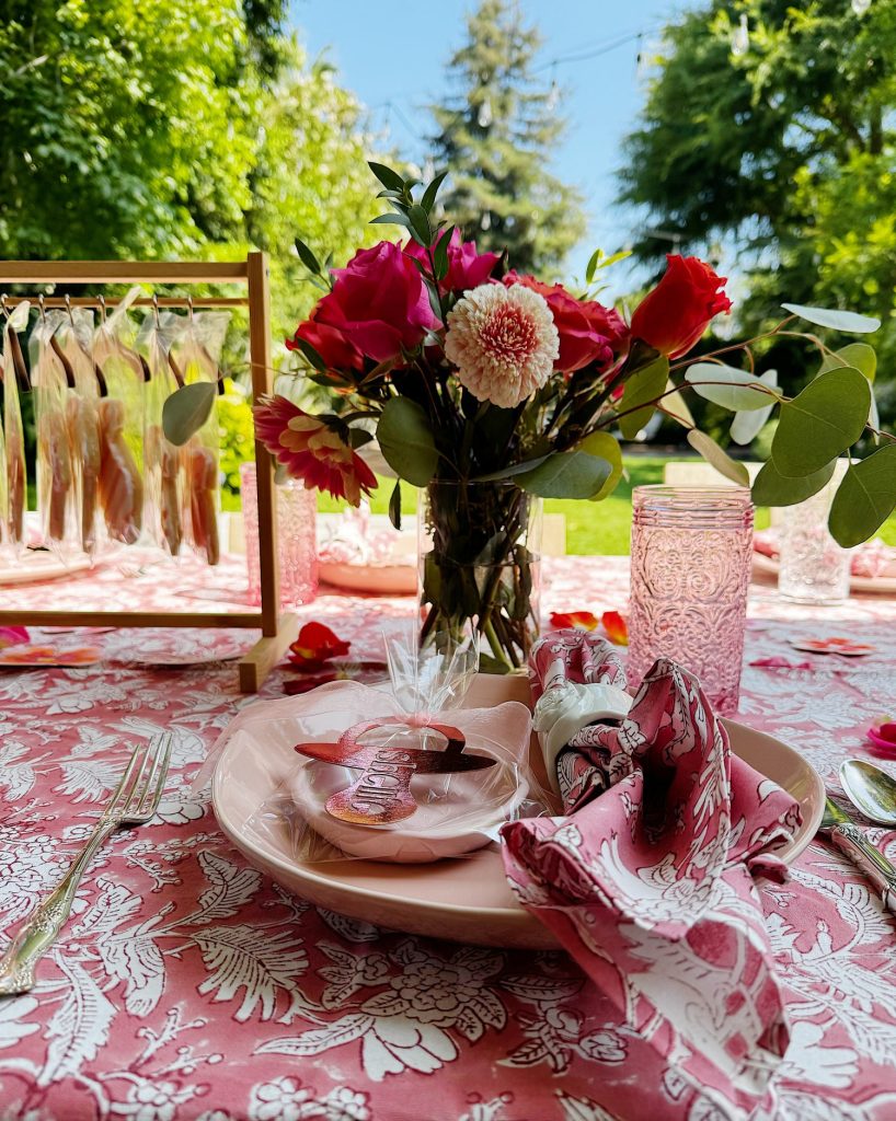 A table set outdoors with pink floral-patterned tablecloth, matching napkins, glassware, a bouquet of red and white flowers, and wrapped cookies at each place setting.